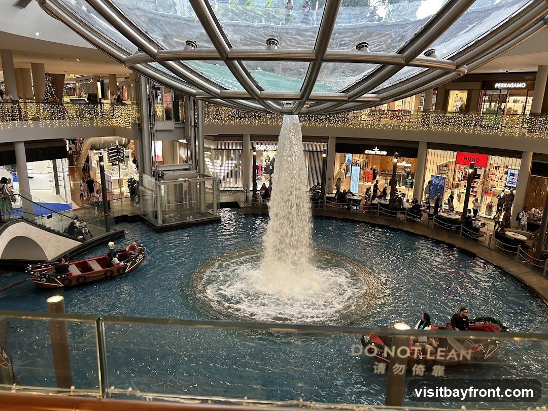 An interior view of a large, multi-level shopping mall featuring a central water feature with a large fountain. Small boats navigate the water, carrying passengers. Shops with visible signage like Ferragamo, Stone Island, and Hugo are present on the upper levels. Decorative lights adorn the balconies.