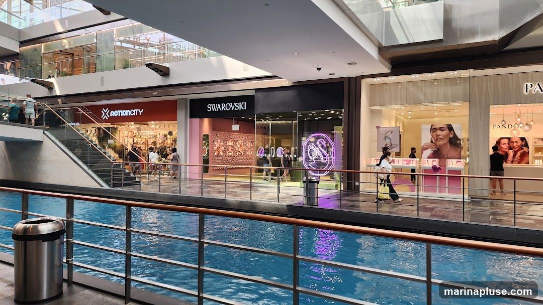 A view of a shopping mall interior with a canal or water feature in the foreground. Several storefronts are visible, including Action City, Swarovski, and Pandora. People are walking around the mall.