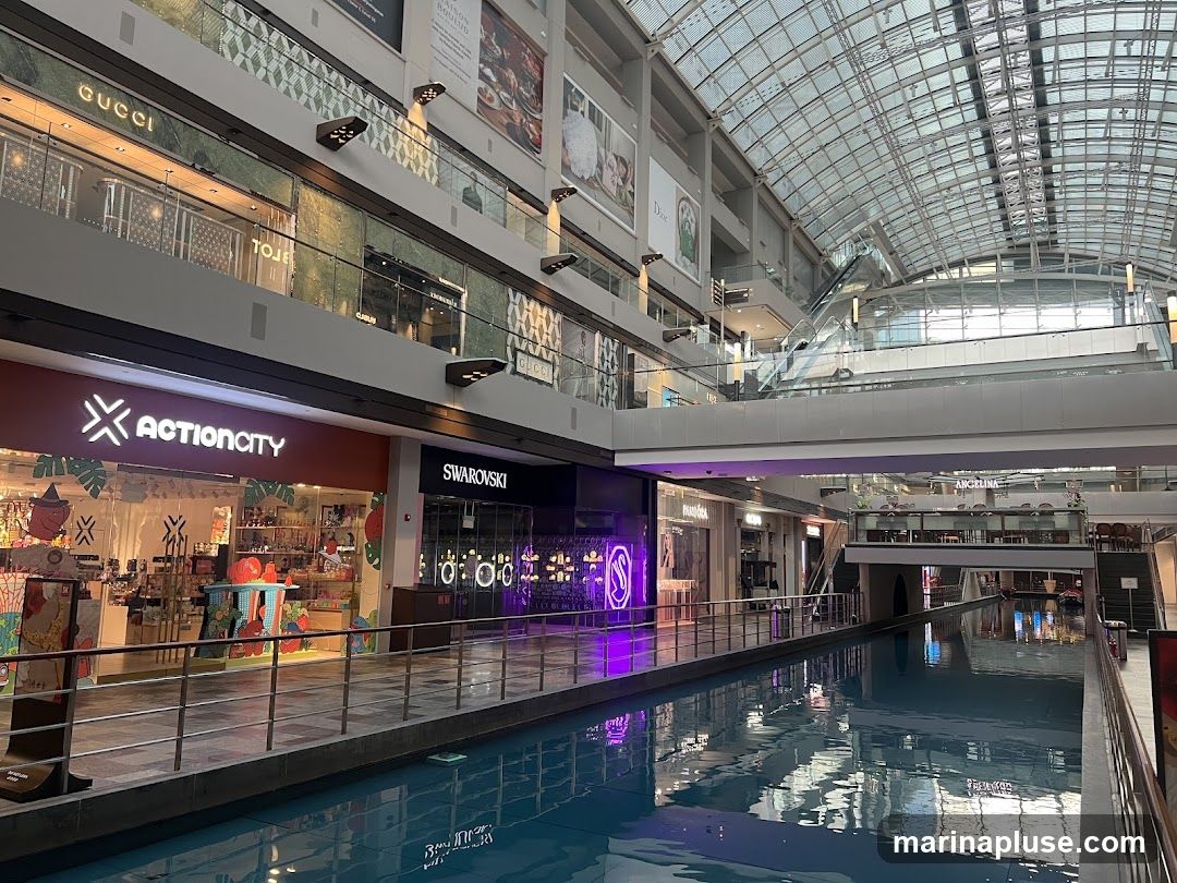 An indoor canal runs through a multi-level shopping mall with various stores visible on the upper floors. Brands like Gucci, Actioncity, and Swarovski are identifiable. The mall features a glass ceiling and escalators.