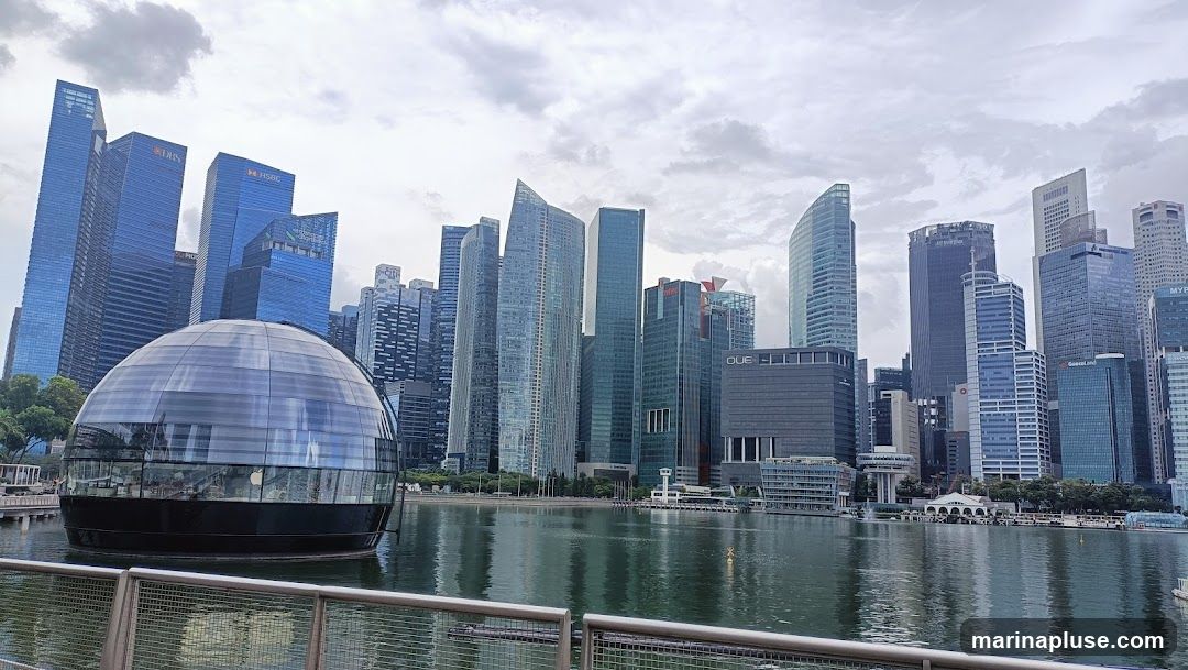 A wide shot of the Marina Bay skyline in Singapore, featuring numerous modern skyscrapers. In the foreground, a distinctive spherical glass building, identified as an Apple Store by its logo, sits on the water. The cloudy sky is reflected in the water along with the buildings.