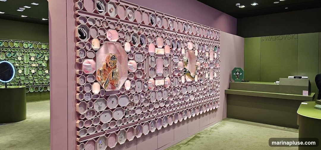 The interior of a Swarovski store with a large display wall featuring jewelry. The wall is pink and has numerous hexagonal and octagonal compartments, many of which contain necklaces, earrings, and rings. A golden statue of a person holding a mirror is displayed prominently in one of the larger compartments. To the left, another display wall is visible, filled with green-toned items. A counter area with the Swarovski logo is seen on the right, along with a Mickey Mouse 100th anniversary sign.