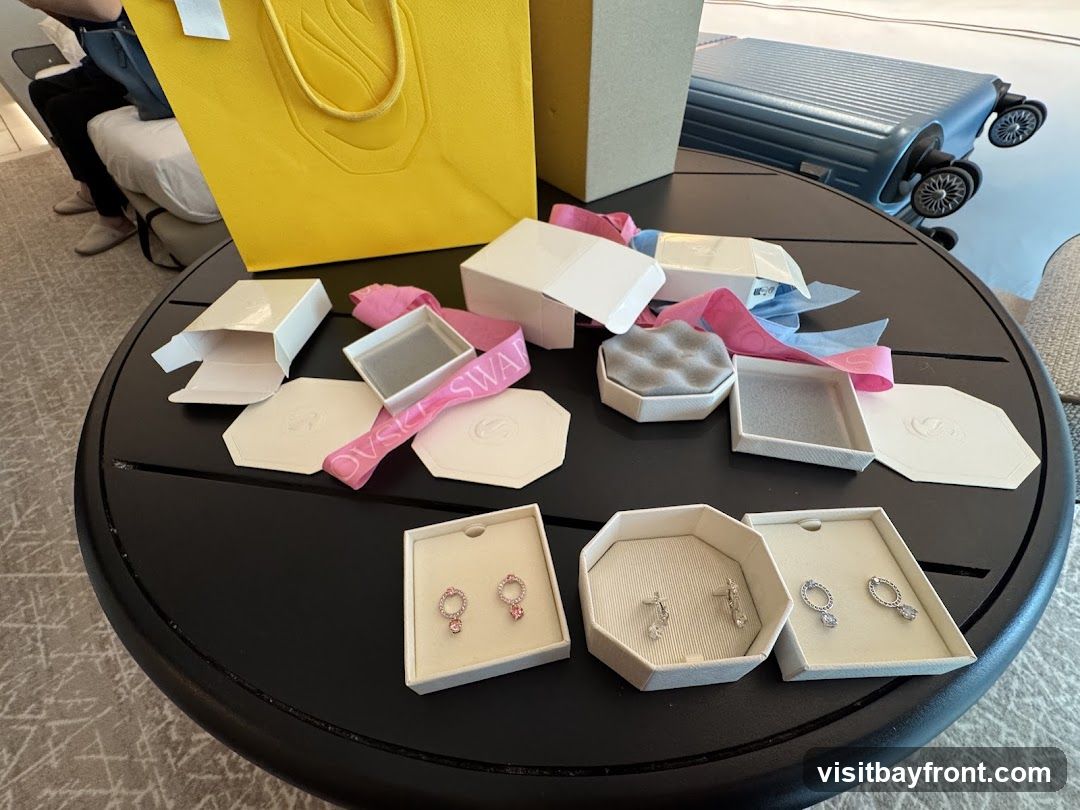 A collection of opened jewelry boxes, ribbons, and gift bags are spread across a round black table. Three pairs of earrings are displayed in their boxes: one pair with pink stones, one pair with clear stones, and another pair with clear stones. A yellow shopping bag and a blue suitcase are visible in the background.