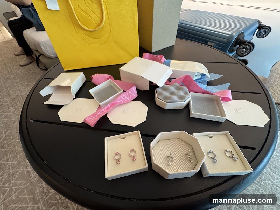 A collection of opened jewelry boxes, ribbons, and gift bags are spread across a round black table. Three pairs of earrings are displayed in their boxes: one pair with pink stones, one pair with clear stones, and another pair with clear stones. A yellow shopping bag and a blue suitcase are visible in the background.