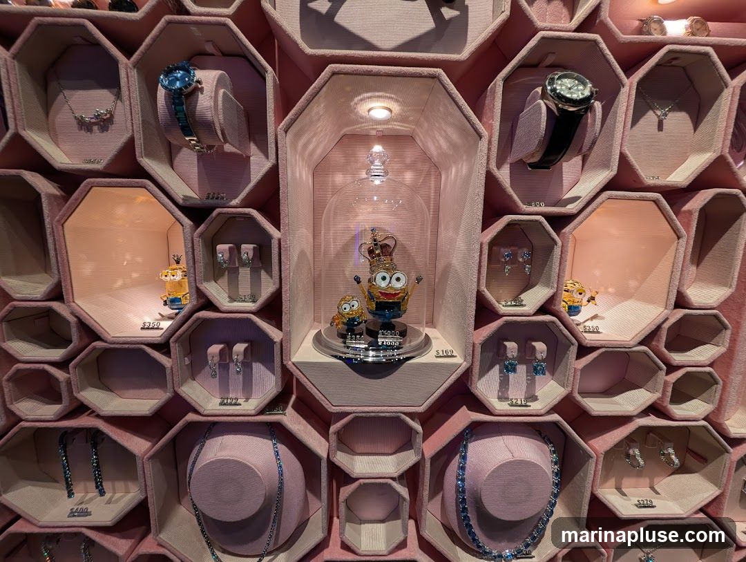 A display case filled with jewelry and figurines. In the center, a Minion character wearing a crown is displayed under a glass dome, priced at $169. To the left and right of the central display are smaller hexagonal compartments showcasing various jewelry items such as necklaces, earrings, and watches. Some compartments also feature smaller Minion figurines, one priced at $350. The display is organized in a honeycomb-like pattern with pink velvet lining.
