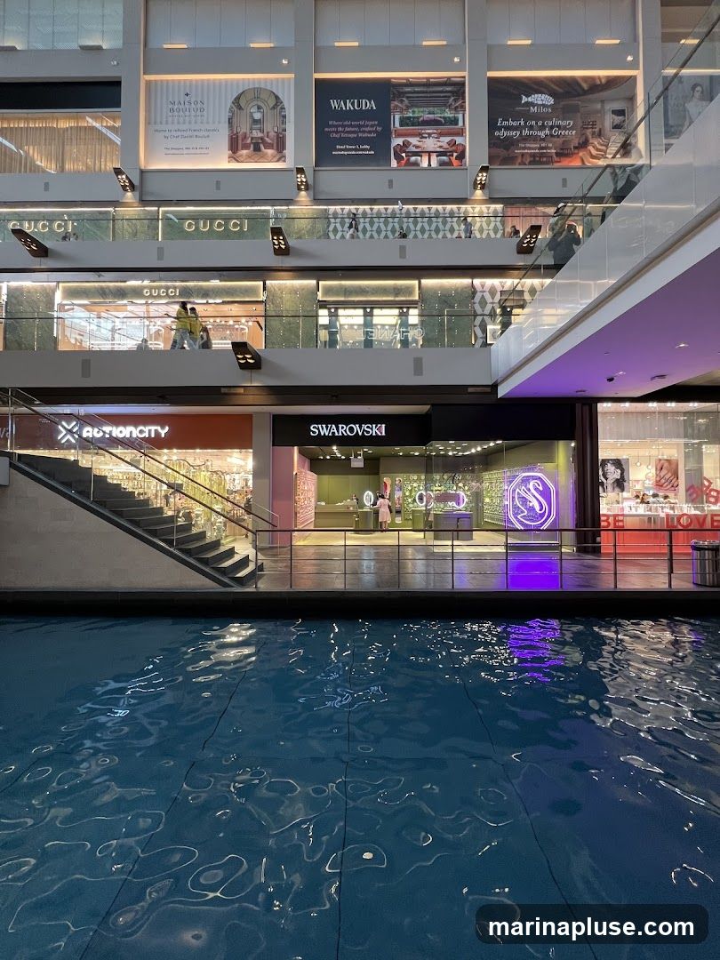 An indoor shopping mall with multiple levels of stores and escalators. A large body of water with rippling reflections is in the foreground. Stores visible include Gucci, Actioncity, and Swarovski. Advertisements for restaurants and brands are displayed on upper levels.