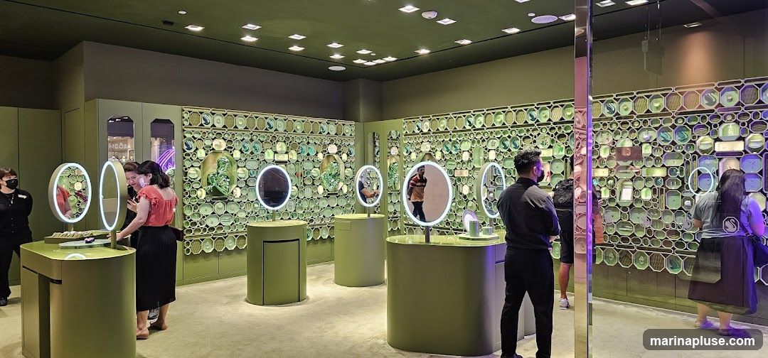 A brightly lit jewelry store with green walls and displays. Several customers are browsing the merchandise, which is arranged on shelves and in display cases. Some of the displays feature illuminated mirrors. The walls are decorated with a patterned mosaic of geometric shapes and what appear to be jewelry pieces.