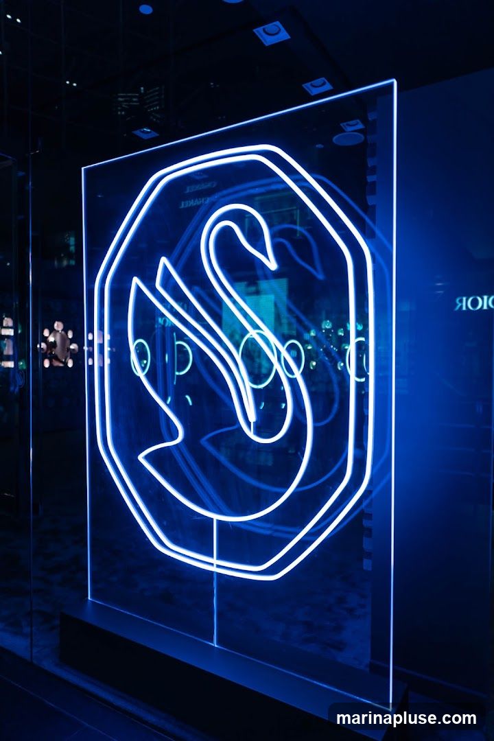 A large, clear acrylic panel displays a swan logo outlined in bright white neon lights. The logo is set within an octagonal frame. The background is dark, with reflections of lights and possibly other displays visible on the acrylic and in the surrounding environment. The overall aesthetic is modern and illuminated, with a strong blue hue dominating the scene.