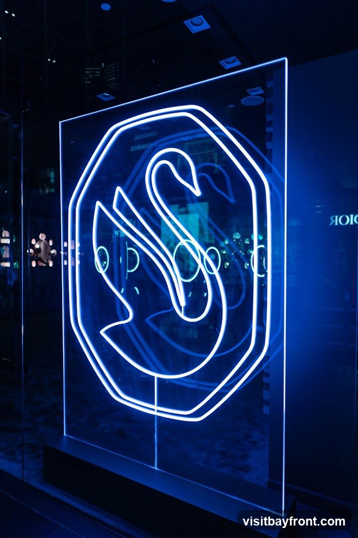 A large, clear acrylic panel displays a swan logo outlined in bright white neon lights. The logo is set within an octagonal frame. The background is dark, with reflections of lights and possibly other displays visible on the acrylic and in the surrounding environment. The overall aesthetic is modern and illuminated, with a strong blue hue dominating the scene.