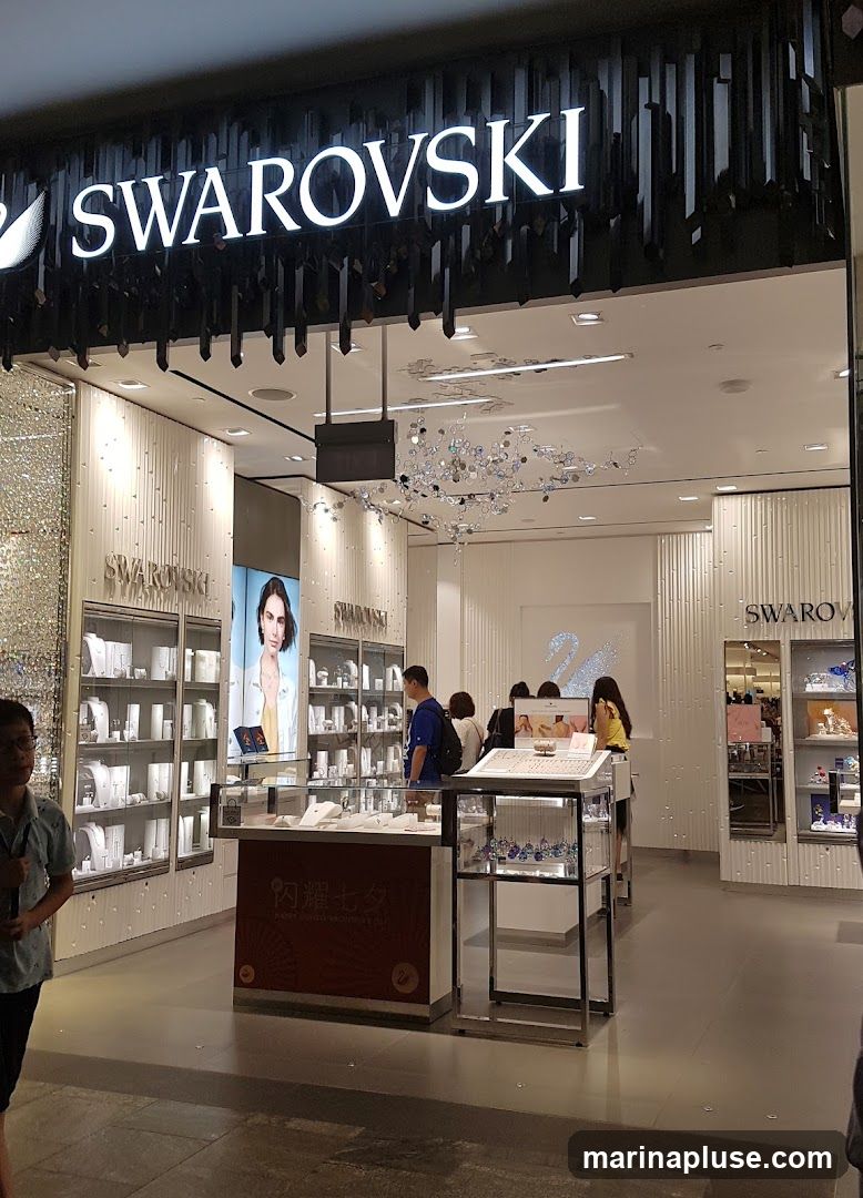 The exterior of a Swarovski store with the brand name prominently displayed above the entrance. The store features illuminated displays of jewelry and accessories behind glass cases. Several customers are browsing inside.