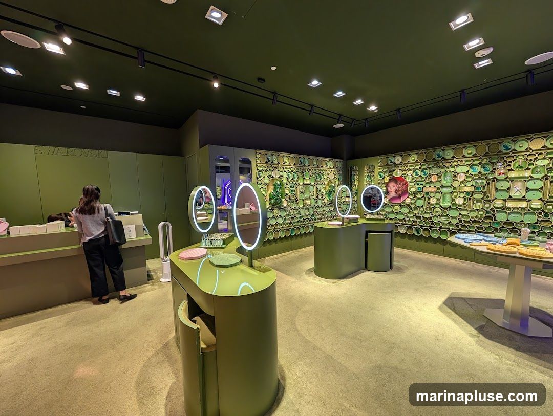 The interior of a Swarovski store with a customer at the counter. The store is decorated in shades of green, with display walls showcasing various jewelry and accessories. There are illuminated mirrors on display tables, and a large wall display with numerous green-framed items.