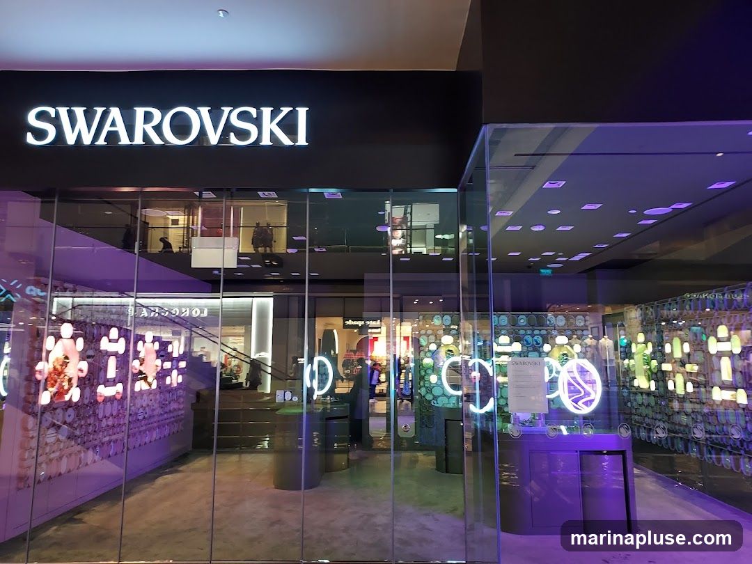 The exterior of a Swarovski store with large glass windows and doors. The prominent "SWAROVSKI" logo is displayed in white illuminated letters above the entrance. Inside, the store is brightly lit with purple and white lights, showcasing displays of jewelry and accessories. Reflections of the mall's interior are visible on the glass.
