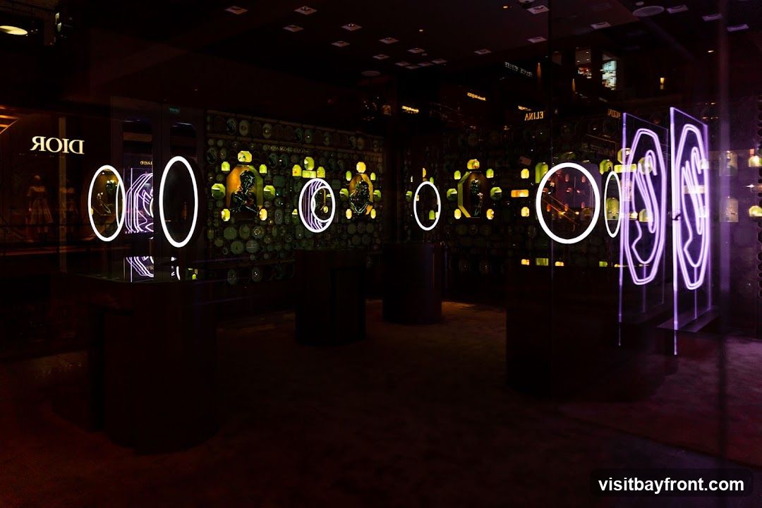 The interior of a Dior store is dimly lit with neon accents. The walls are decorated with a mosaic of circular and square objects, possibly watches or jewelry, illuminated by yellow lights. Several display cases with white and purple neon rings showcase products. A mannequin in a dress is visible through the window on the left.
