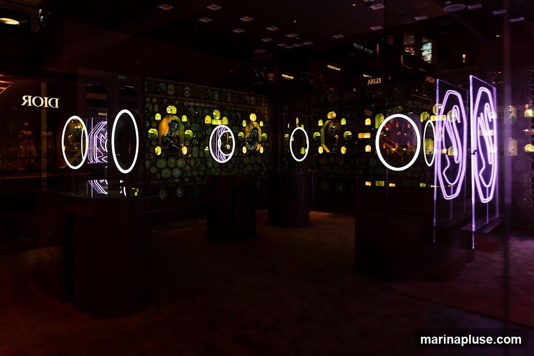 The interior of a Dior store is dimly lit with neon accents. The walls are decorated with a mosaic of circular and square objects, possibly watches or jewelry, illuminated by yellow lights. Several display cases with white and purple neon rings showcase products. A mannequin in a dress is visible through the window on the left.