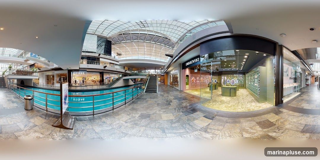 A panoramic view of an indoor shopping mall with a canal-like water feature. Several storefronts are visible, including Guess, Swarovski, and Pandora. The mall has a modern design with a large glass roof and multiple levels. People are seen browsing in the stores and walking through the mall.