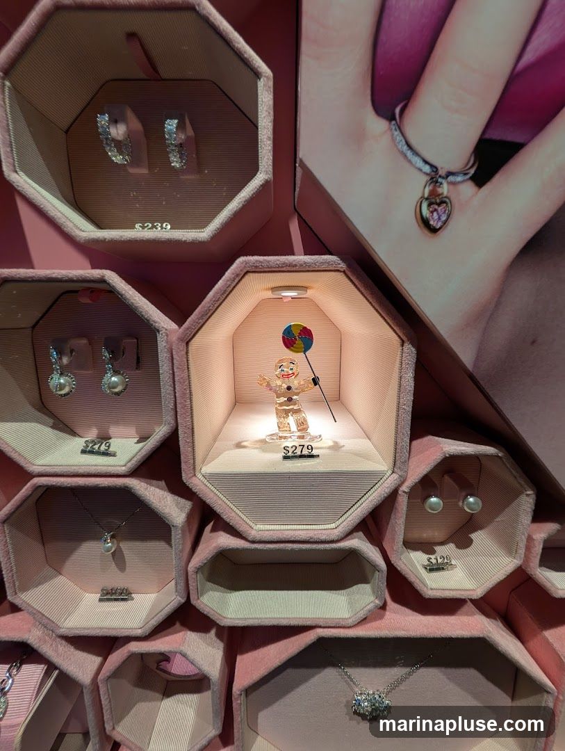 A display case filled with various jewelry items, including earrings, necklaces, and rings. The display is organized in hexagonal compartments lined with pink velvet. Some items have price tags: $239, $279, $169, and $129. A decorative figurine of a person holding a lollipop is also displayed. A large promotional image of a hand wearing a ring with a heart-shaped pendant is visible in the background.