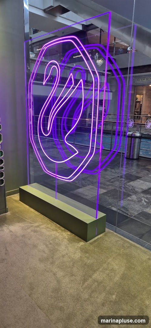A large, illuminated neon sign in the shape of a swan within an octagon is displayed on a clear panel. The neon light is a vibrant purple. The display is set on a dark green base and is positioned in what appears to be a public indoor space, possibly a mall, with a carpeted floor and tiled areas visible. In the background, other shops and people can be seen, including a sign that reads "CLUB MO".