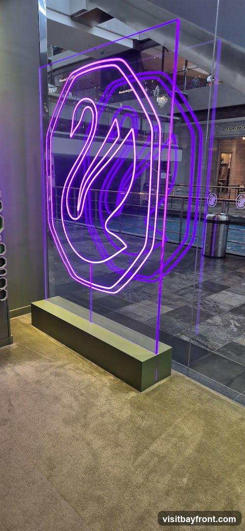 A large, illuminated neon sign in the shape of a swan within an octagon is displayed on a clear panel. The neon light is a vibrant purple. The display is set on a dark green base and is positioned in what appears to be a public indoor space, possibly a mall, with a carpeted floor and tiled areas visible. In the background, other shops and people can be seen, including a sign that reads "CLUB MO".