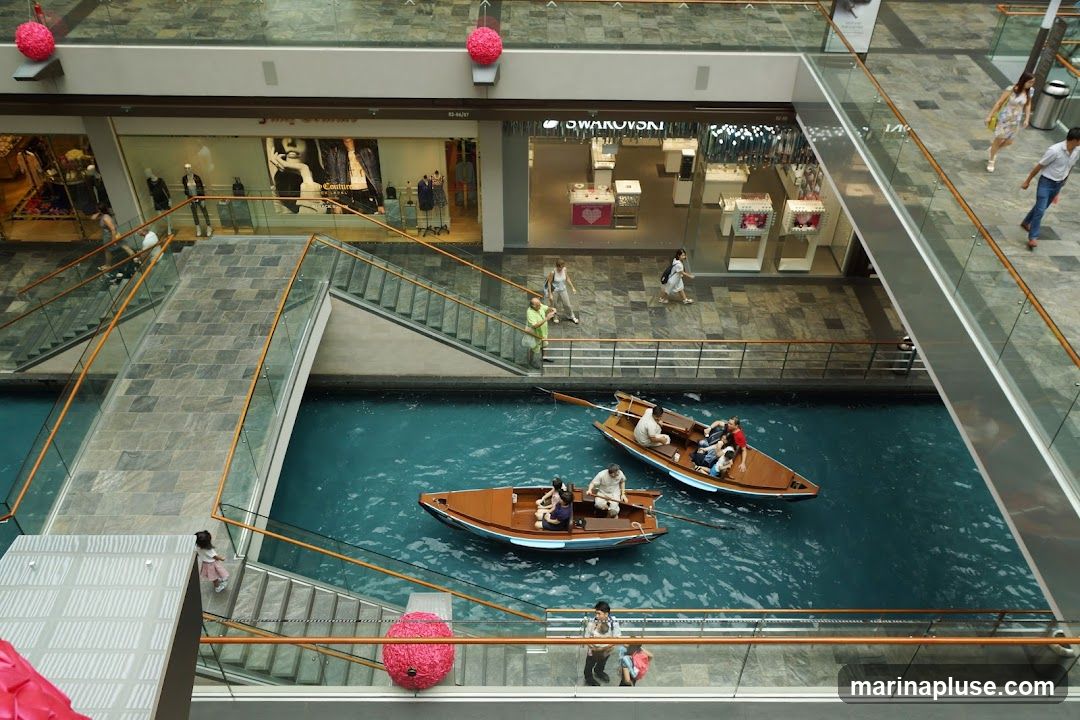 An aerial view of a shopping mall with a waterway running through it. People are seen riding in small wooden boats on the blue water. Escalators and walkways are visible above, with various shops lining the upper levels. One shop has a prominent "SWAROVSKI" sign.