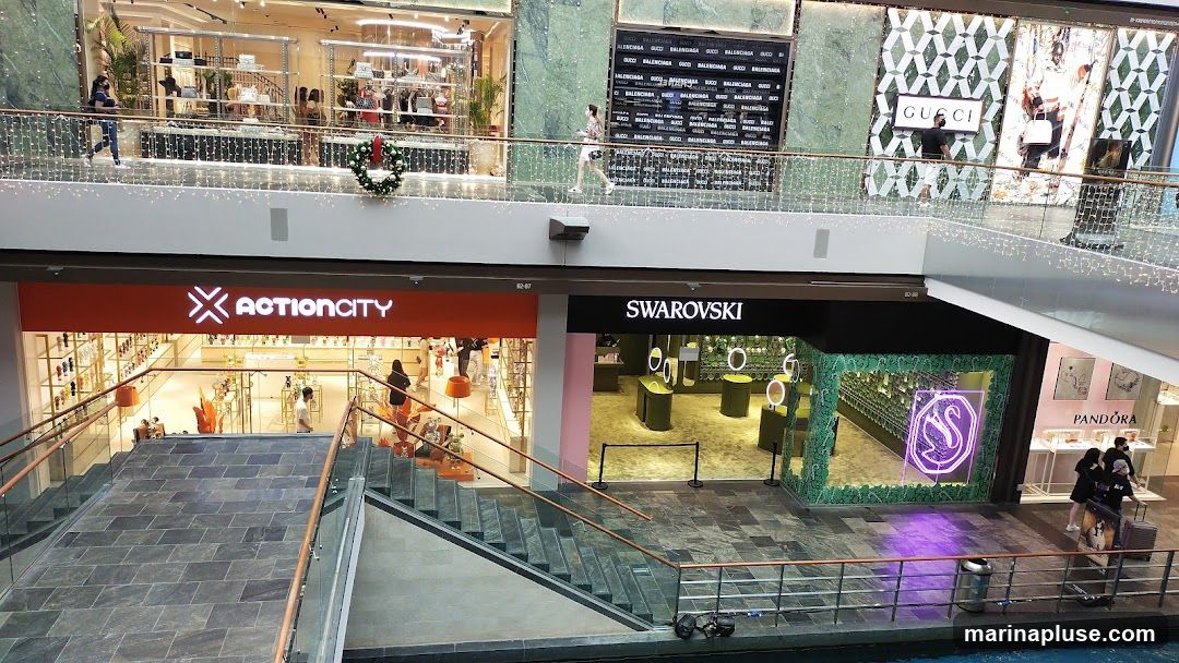 A view of a mall interior showing multiple storefronts on different levels. The foreground features a wide staircase leading down. On the lower level, the storefronts for ActionCity and Swarovski are visible, with their respective signage. Further back, on the upper level, storefronts for Gucci and Pandora are also visible. People are seen walking around the mall.