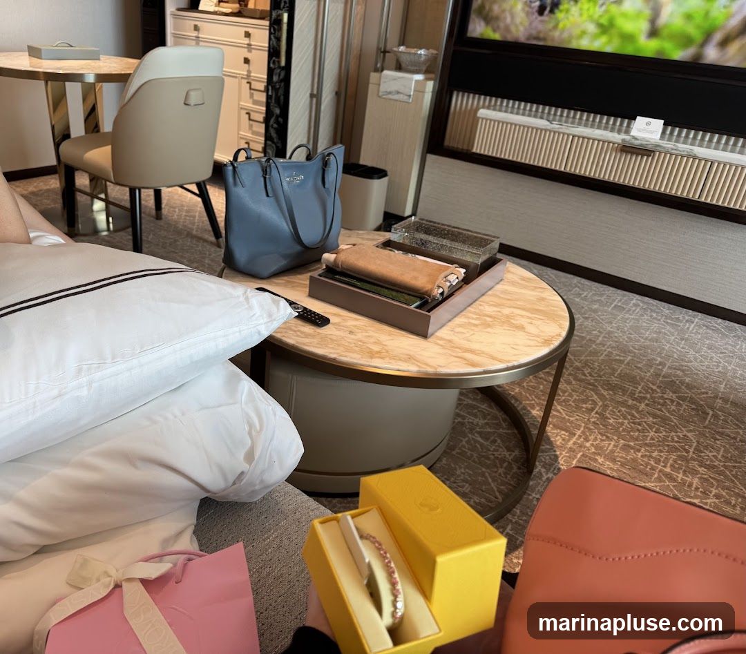 A hotel room is depicted with a bed, a coffee table, and a chair. On the coffee table, there is a blue handbag, a tray with folded items, and a decorative box. In the foreground, a pink gift bag and a yellow jewelry box containing a bracelet are visible. A pink handbag is also present. The room appears to be well-appointed, with a television screen visible in the background.