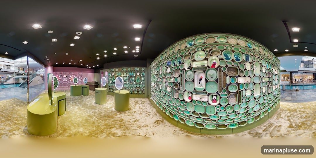 The interior of a store with a large, decorative wall displaying various items in hexagonal and rectangular compartments. The compartments contain jewelry, figurines, and other decorative objects, many in shades of green and blue. In the foreground, there are green display stands with mirrors. The floor is covered in a textured, light-colored carpet. Through a glass entrance on the left, a shopping mall interior with escalators and other stores is visible. On the right, another store with a "GUESS" sign is visible across a walkway.