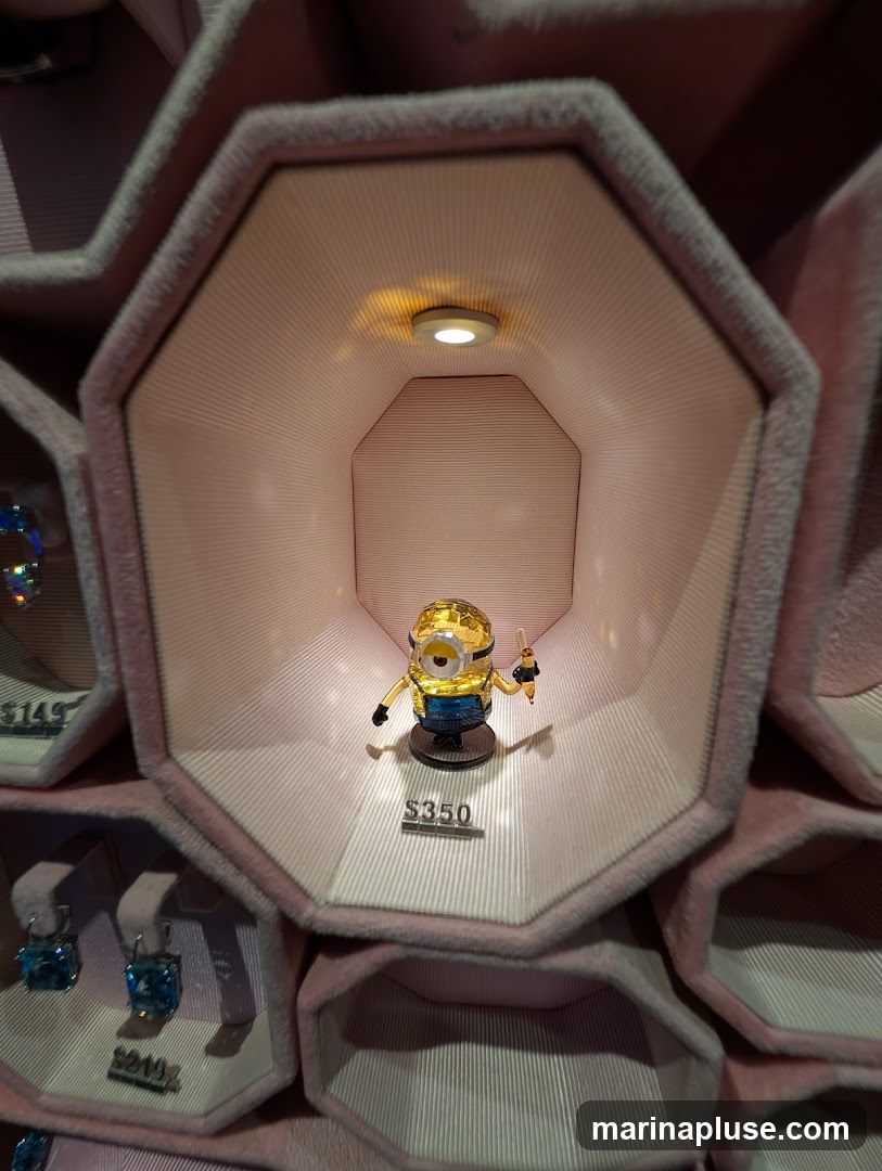 A crystal figurine of a Minion character is displayed in a pink, hexagonal-shaped case with a light illuminating it from above. The figurine is gold and blue and holds a small object in its hand. A price tag of $350 is visible below the figurine. Other display cases with jewelry are partially visible to the left and below, with price tags of $149 and $249.