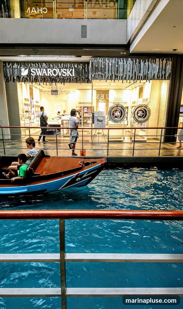 The exterior of a Swarovski store in a mall. The store has a large glass facade with the Swarovski logo prominently displayed above the entrance. Inside, shelves are stocked with merchandise, and customers are browsing. In the foreground, a boat with passengers sails on a canal.