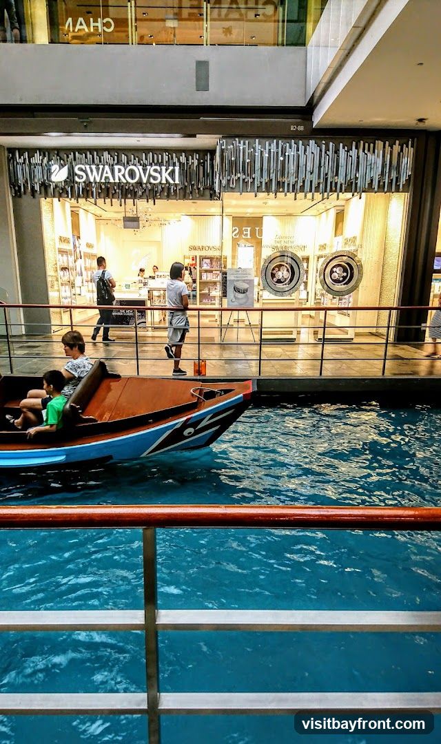 The exterior of a Swarovski store in a mall. The store has a large glass facade with the Swarovski logo prominently displayed above the entrance. Inside, shelves are stocked with merchandise, and customers are browsing. In the foreground, a boat with passengers sails on a canal.