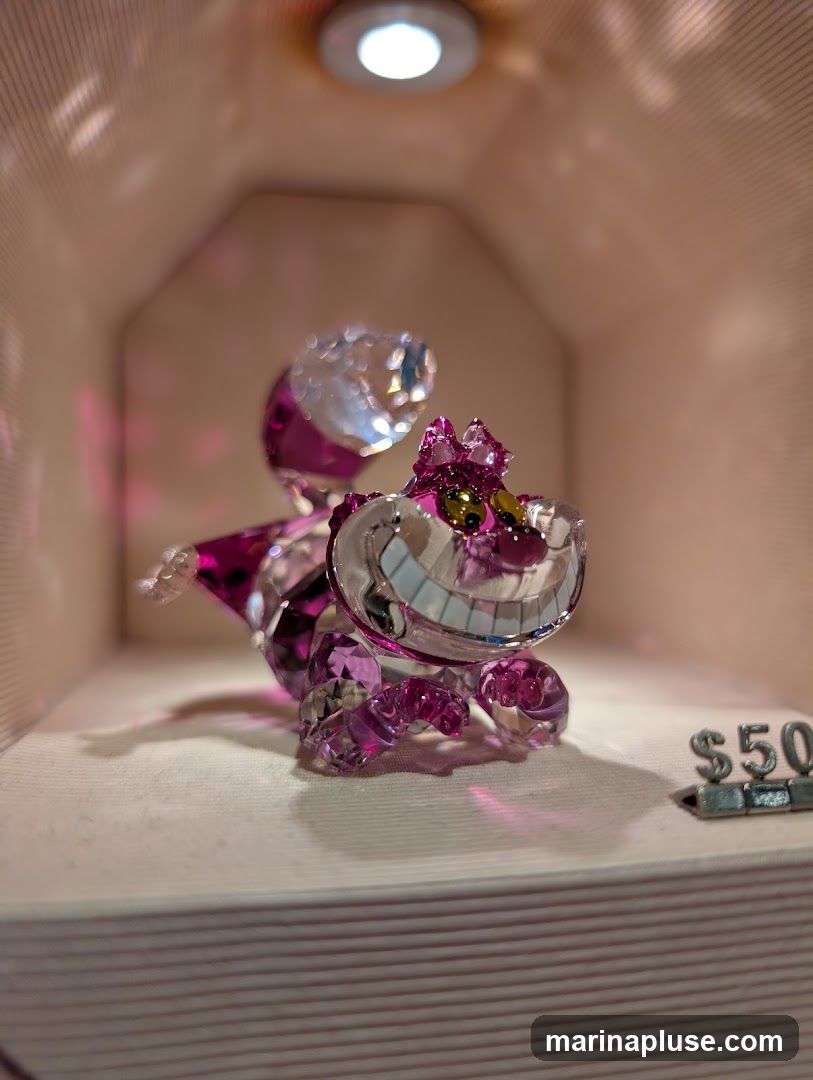 A close-up shot of a sparkling crystal figurine of the Cheshire Cat from Alice in Wonderland. The figurine is predominantly pink and clear, with intricate facets that catch the light. It is displayed on a white surface within a softly lit, pink-toned display case. A small price tag showing '$50' is visible to the right of the figurine.