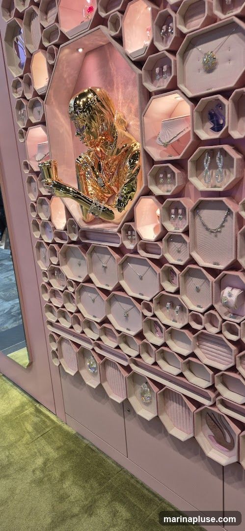 A display case filled with jewelry, featuring a golden sculpture of a person in the center. The display is organized into hexagonal compartments, each showcasing necklaces, earrings, bracelets, and rings. The overall aesthetic is elegant and luxurious, with a pink background and soft lighting.