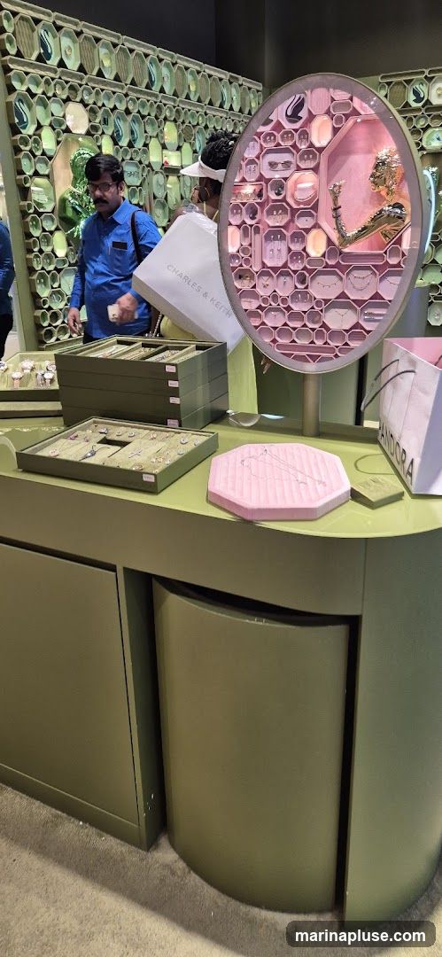A display of jewelry in a store. There are several trays of necklaces and earrings on a green counter. A pink octagonal display with a necklace sketch is in the foreground. In the background, a wall is decorated with hexagonal and oval shapes, some containing swan figurines. Two shoppers are visible, one holding a Charles & Keith shopping bag and another looking at the display. A Pandora shopping bag is also visible.