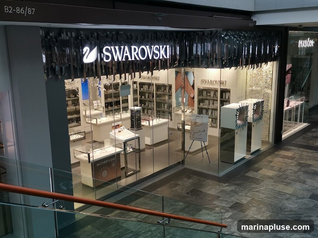 The exterior of a Swarovski jewelry store. The store has a large glass facade displaying various jewelry items in display cases. The prominent Swarovski logo with a swan is visible above the entrance. Adjacent to the Swarovski store is a Pandora store.