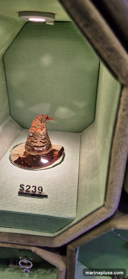 A decorative glass witch hat with an iridescent finish is displayed on a light green velvet-lined platform inside a display case. A price tag showing '$239' is placed in front of the hat. The display case has a hexagonal shape and is also lined with green fabric. A small, jeweled object, possibly a charm or pendant, is visible at the bottom of the frame.