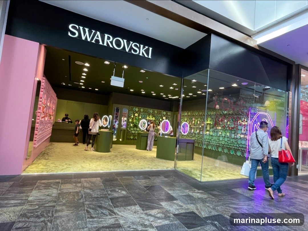 The exterior of a Swarovski store with the brand name prominently displayed above the entrance. The store features a modern design with large glass windows showcasing displays of jewelry and accessories. Inside, customers are browsing the merchandise.