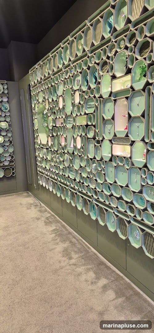 A wall display with numerous hexagonal and octagonal compartments, each showcasing jewelry items such as necklaces and pendants. The compartments are lined with a light green material, and some contain small spotlights illuminating the displayed pieces. The overall aesthetic is modern and elegant, with a focus on presenting the jewelry in an organized and visually appealing manner. The floor is carpeted in a neutral tone.