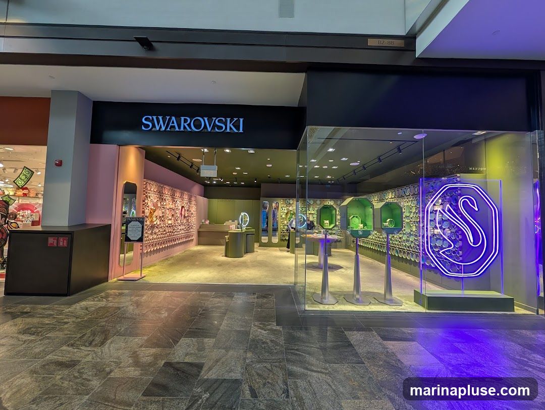 The exterior of a Swarovski store with the brand name prominently displayed above the entrance. The storefront features a large glass window showcasing various jewelry displays and a neon swan logo. Inside, merchandise is arranged on shelves and display stands.