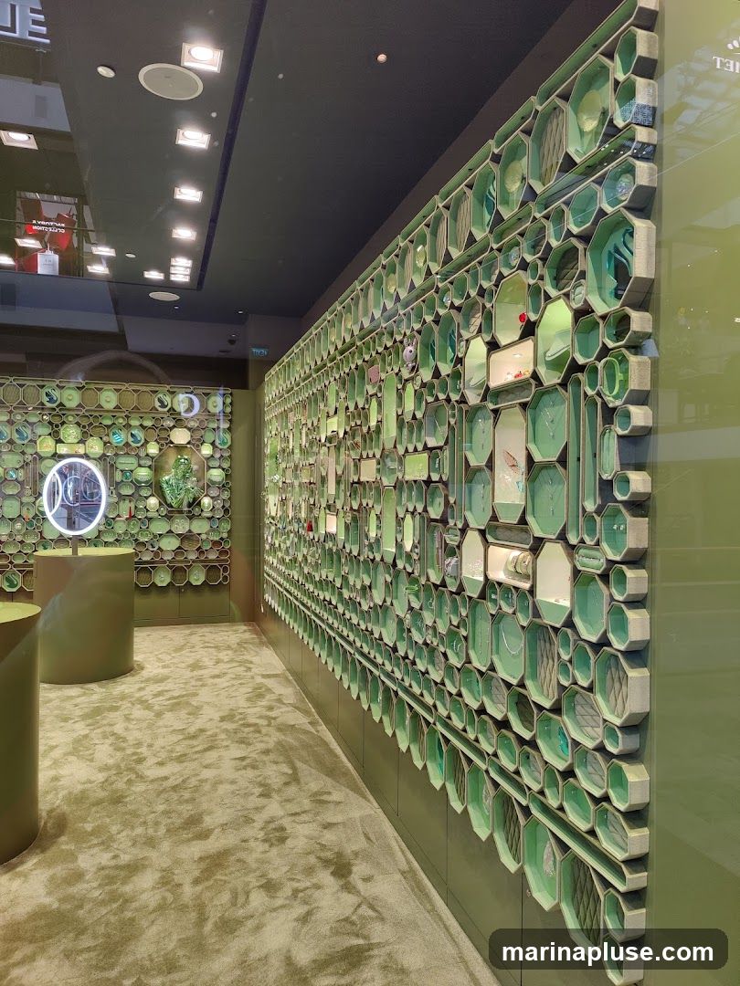 The interior of a store with a large, hexagonal-patterned display wall filled with various items. The wall is predominantly green and grey, with some items appearing to be jewelry or decorative objects. The floor is carpeted in a textured green. Two cylindrical display stands are visible in the foreground. In the background, another display wall with circular elements and a lit mirror can be seen. The ceiling is dark with recessed lighting.