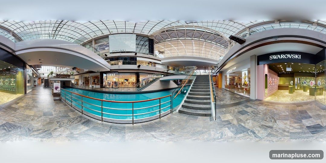 A panoramic view of the interior of a modern shopping mall. A canal with turquoise water runs through the center, bordered by railings. Escalators and staircases lead to upper levels. Various shops are visible, including Swarovski and Lancaster, with their storefronts displaying merchandise. The mall features a large, arched glass ceiling allowing natural light to illuminate the space.