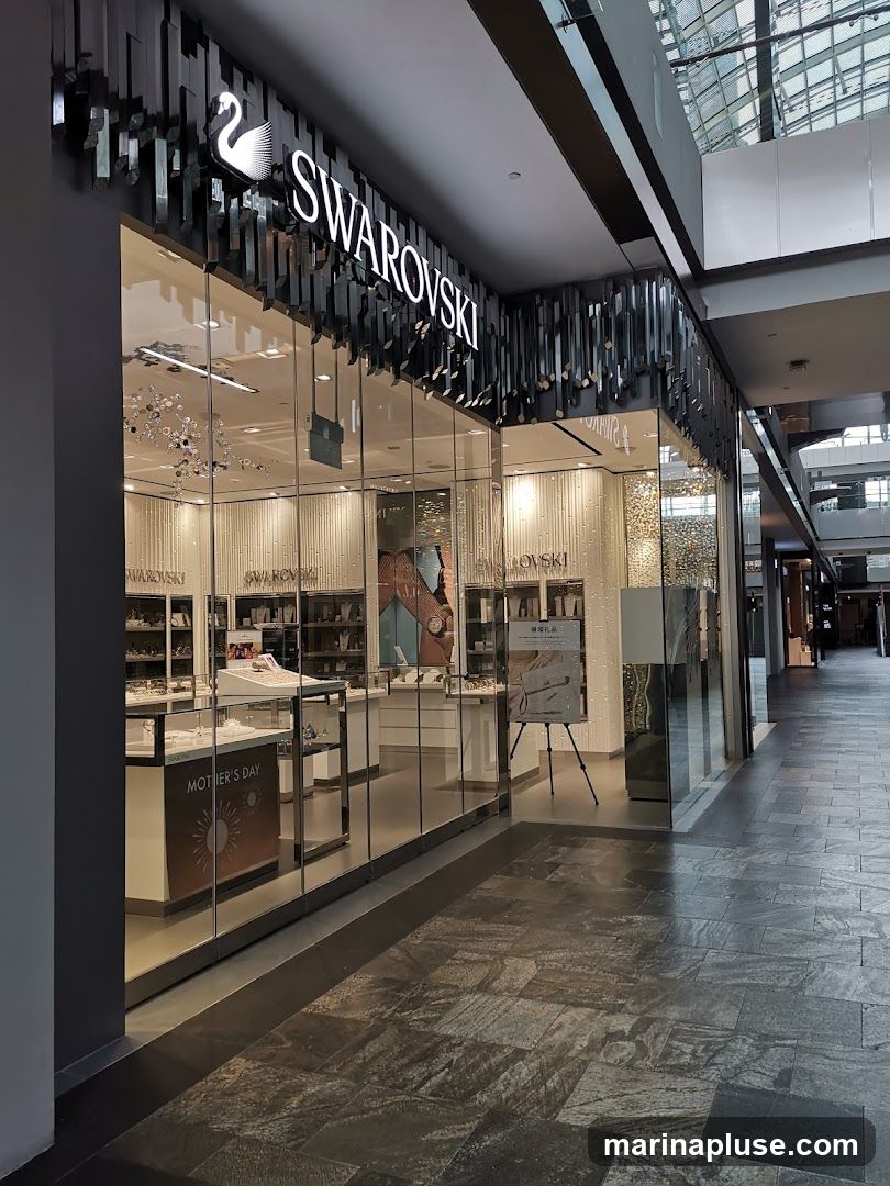 The exterior of a Swarovski store with a large glass facade. The store's name and logo are prominently displayed above the entrance. Inside, display cases showcase jewelry and accessories. A "Mother's Day" promotional display is visible.