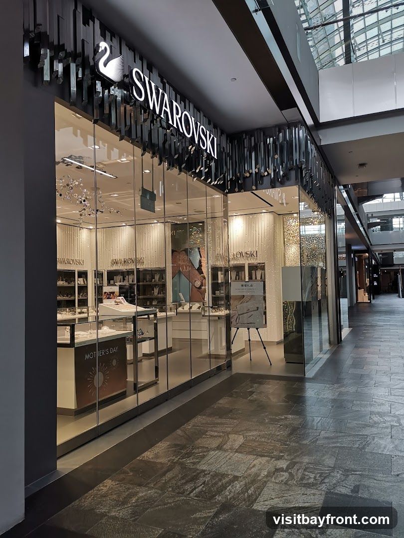 The exterior of a Swarovski store with a large glass facade. The store's name and logo are prominently displayed above the entrance. Inside, display cases showcase jewelry and accessories. A "Mother's Day" promotional display is visible.