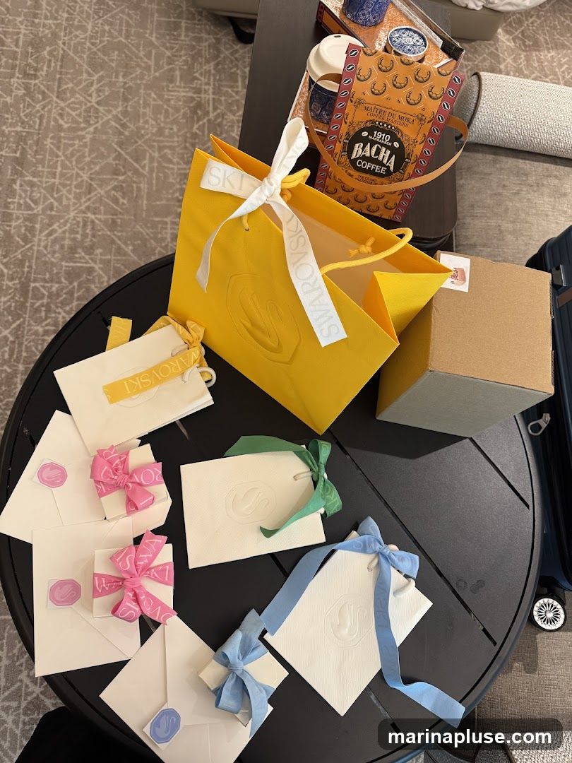 A collection of yellow and white Swarovski shopping bags and small gift boxes tied with ribbons in pink, green, and blue. A Bacha Coffee bag is also visible, along with a cardboard box and a suitcase.