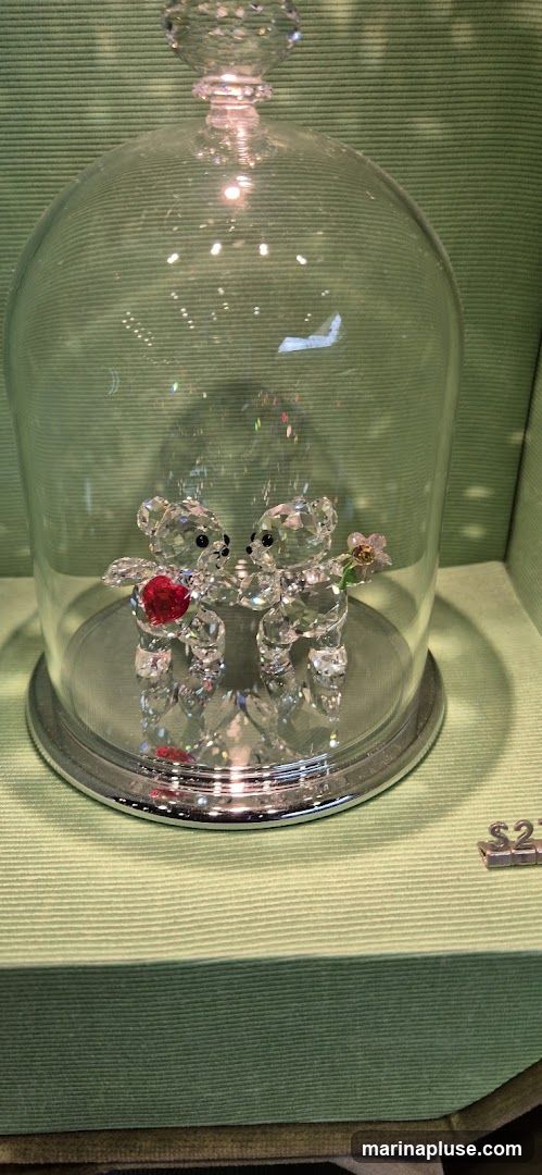 A close-up shot of two clear crystal teddy bears displayed under a glass dome. The teddy bear on the left holds a red heart, and the one on the right holds a small pink flower. The display sits on a textured green surface. A price tag reading '$27' is visible to the right of the dome.