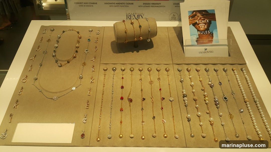 A display of Swarovski Remix Collection jewelry, featuring necklaces and bracelets with various charms and beads. The display includes alphabet charms, pearl strands, and other decorative elements. A promotional booklet with the text "Remix the Rules" is also visible.