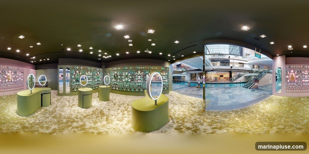A panoramic view of a brightly lit jewelry store interior. The store features a modern design with green display stands and oval mirrors with illuminated rims. The walls are adorned with intricate displays of jewelry, some in pink and others in green. The floor is covered in a textured, light-colored carpet. Through a large glass opening, a shopping mall atrium is visible, with escalators, shops like Chanel and Guess, and a central water feature.