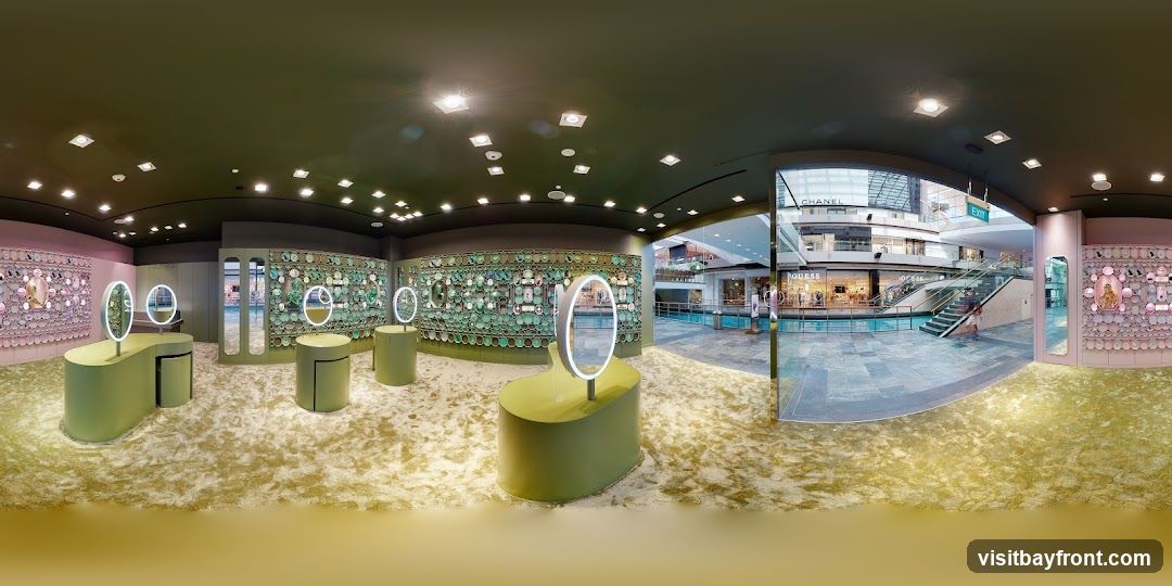 A panoramic view of a brightly lit jewelry store interior. The store features a modern design with green display stands and oval mirrors with illuminated rims. The walls are adorned with intricate displays of jewelry, some in pink and others in green. The floor is covered in a textured, light-colored carpet. Through a large glass opening, a shopping mall atrium is visible, with escalators, shops like Chanel and Guess, and a central water feature.