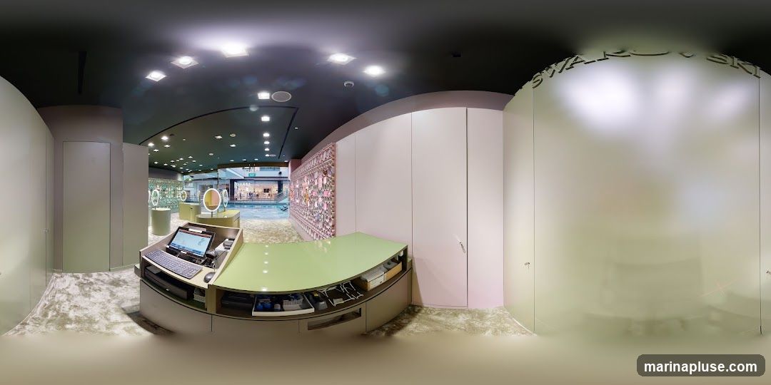 A panoramic view of the interior of a Swarovski store. The image shows a reception desk with a laptop and keyboard, display cases with jewelry, and a large decorative wall panel. In the background, through an open area, other stores and shoppers can be seen. The store has a modern and elegant design with soft lighting. The Swarovski logo is visible on a frosted glass panel on the right side of the image.