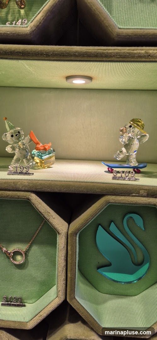 A display case featuring several crystal figurines and jewelry items. In the upper section, a crystal bear wearing a party hat holds a gift, priced at $199. Next to it, another crystal bear rides a skateboard, priced at $169. Above these, a glimpse of another item is visible with a price of $149. The lower section shows a rose gold necklace with a circular pendant, priced at $129, and a teal swan silhouette.