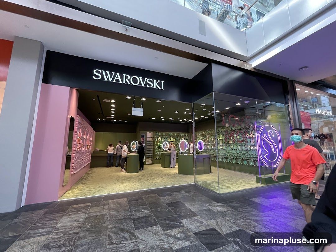 The exterior of a Swarovski store in a mall. The store has a large black sign with "SWAROVSKI" in white lettering above the entrance. The storefront features large glass windows displaying jewelry and a neon swan logo. Several people are visible inside the store.