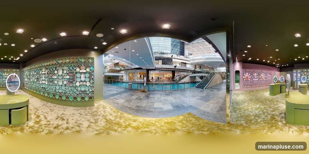 The image shows the interior of a retail store, possibly a jewelry or accessory shop. The walls are decorated with intricate patterns of circular and rectangular elements in shades of green and pink. There are display stands with mirrors and products. In the background, through a large glass facade, a shopping mall interior is visible with other stores, including one with a prominent "GUESS" sign. The floor is covered with a textured carpet.