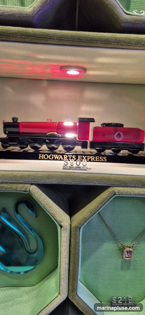 A display case featuring a model of the Hogwarts Express train, illuminated by red lights. Below the train, a sign reads "HOGWARTS EXPRESS" with a price of "$900". To the sides, jewelry is displayed in green velvet boxes. One box contains a swan-shaped pendant, and another holds a necklace with a square-cut gemstone, priced at "$219".